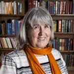 Carol Malleson, an ILS Case Manager from Cardiff, Glamorgan, stands in front of a bookshelf, wearing an orange scarf which complements her grey hair.