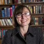 Janine Brooks, an ILS Case Manager based in Brighton, East Sussex, sits in front of a bookcase wearing glasses and a black shirt.
